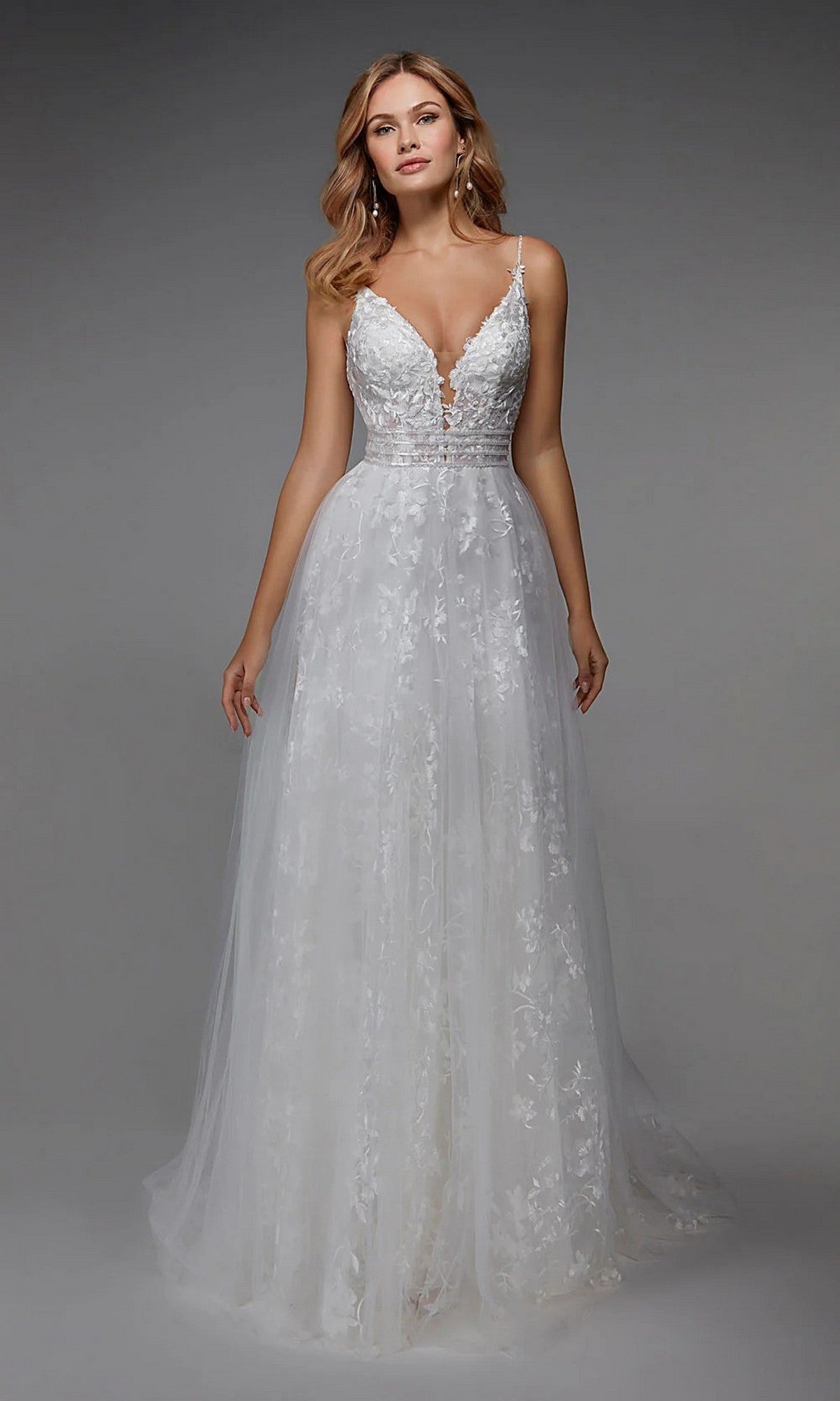 A woman wears the Alyce 7036 long white dress, a sleeveless lace gown with floral appliqués, deep V-neckline, and flowing tulle skirt, posed against a plain gray background.